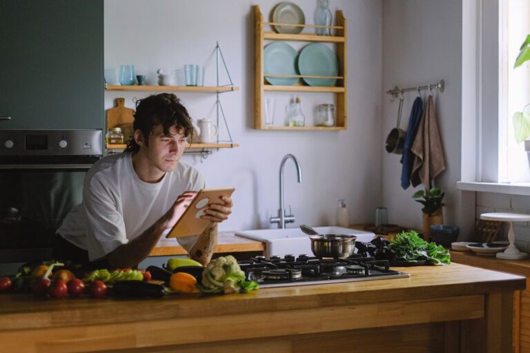 Pessoa utilizando tablet na bancada da cozinha cercada de vegetais frescos para gestão de compras de restaurante.